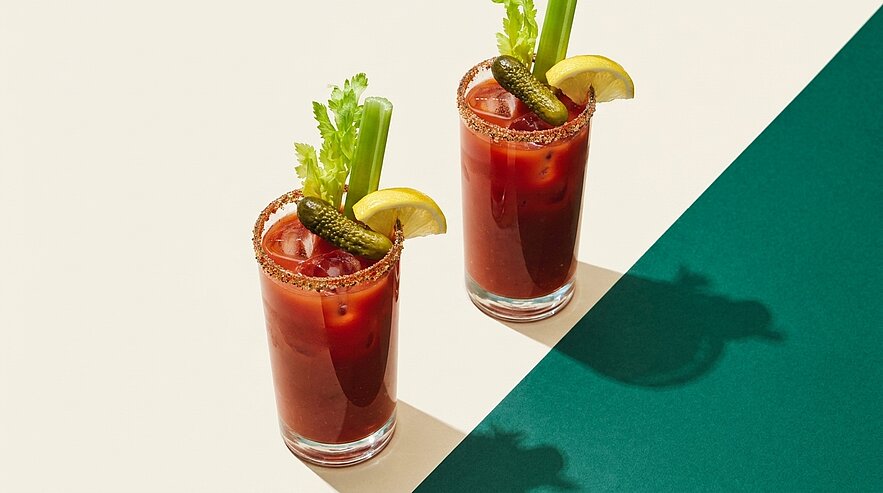 Zwei Bloody-Mary-Cocktails in hohen Gläsern, garniert mit Selleriestangen, Zitronenscheiben, kleinen Gewürzgurken und gewürztem Glasrand.