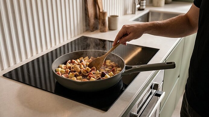 Eine Hand rührt mit einem Holzlöffel gewürfelte Kartoffeln in einer Pfanne auf dem Herd, es steigt Dampf auf.