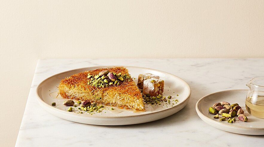 Ein Stück Baklava auf einer runden, hellen Servierplatte, dekoriert mit gehackten Pistazien. Neben dem Kuchen liegt ein kleiner Bissen und eine separate Schale mit weiteren Pistazien sowie einer transparenten Flüssigkeit. Der Hintergrund ist schlicht und unauffällig.