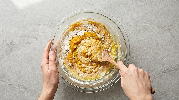Zwei Hände rühren mit einem Holzlöffel in einer Glasschüssel einen zähflüssigen Kuchenteig.