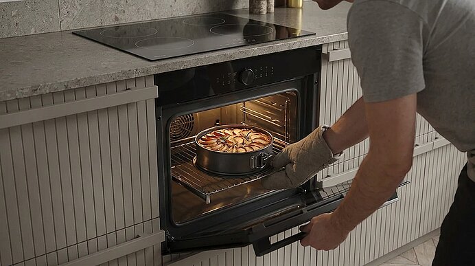 Eine Person mit Ofenhandschuhen schiebt eine runde Obsttorte in einen eingebauten Backofen.