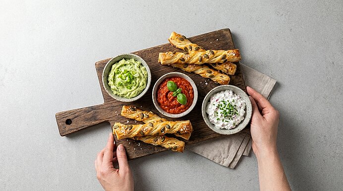 Hand hält Holzbrett mit knusprigen Blätterteigstangen und drei Schälchen Dips: Guacamole, Tomatensalsa mit Basilikum und Kräuterjoghurt.