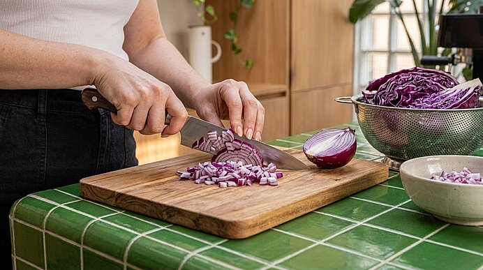 Hände schneiden mit einem Messer eine rote Zwiebel auf einem Holzbrett; gewürfelte Zwiebelstücke liegen daneben, im Hintergrund Rotkohl im Sieb.