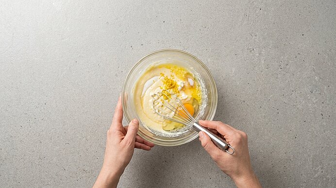 Zwei Hände verrühren Quark, Eier, Zucker, Zitronenabrieb und Butter mit einem Schneebesen in einer Glasschüssel.