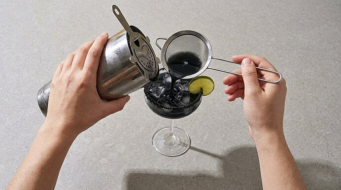 Zwei Hände seihen schwarze Margarita aus einem Shaker durch ein Sieb in ein Glas mit Eiswürfeln.