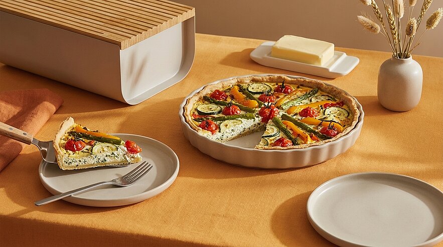 Runde Gemüsequiche mit Zucchinistreifen und Kirschtomaten in einer Backform; eine Scheibe auf einem Teller mit Gabel serviert.