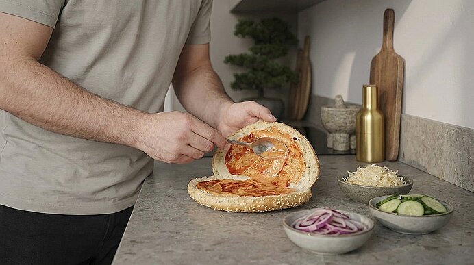 Hände streichen Tomatensoße auf ein aufgeschnittenes Sesambrötchen; daneben Schalen mit roten Zwiebeln, Gurkenscheiben und geriebenem Käse.