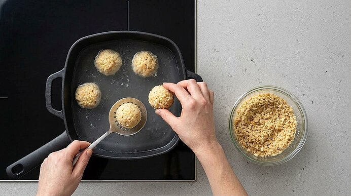 Zwei Hände legen geformte Riebelmais-Knödel mit einem Schaumlöffel in einen Topf mit leicht siedendem Wasser.