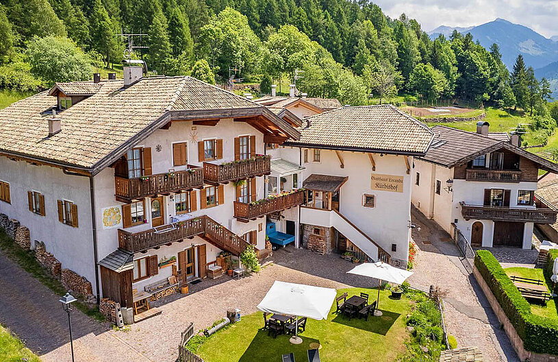 Alpines Gästehaus-Ensemble mit weiß verputzten Gebäuden und Holzbalustraden, Innenhof mit Sitzplätzen und Sonnendach, eingebettet in grüne Wiesen, Nadelwald und dahinter aufragende Berggipfel.