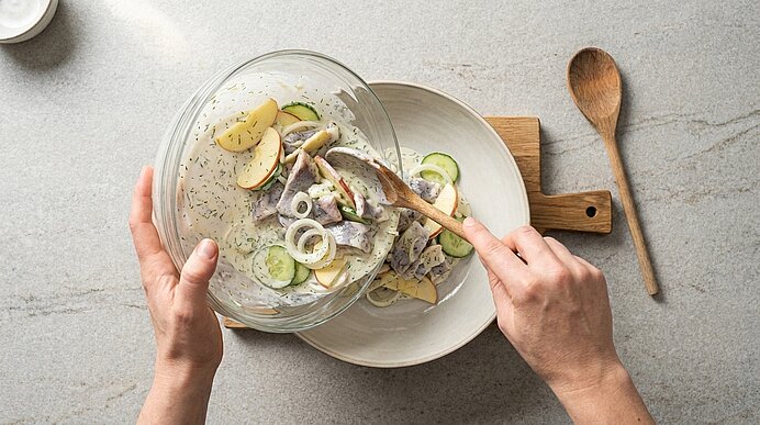 Hände servieren einen cremigen Kartoffel‑Gurken‑Salat mit Zwiebeln und eingelegtem Hering aus einer Glasschüssel in einen Teller.
