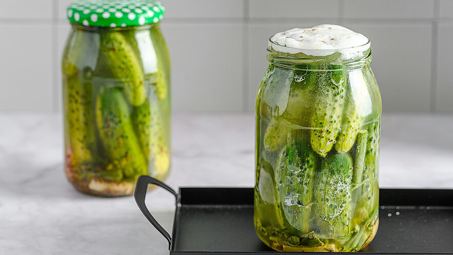 Zwei transparente Gläser mit eingelegten Gurken stehen auf einer Tischoberfläche. Das vordere Glas ist voll mit grünen Gurken und einer schaumigen Flüssigkeit, während das hintere Glas einen grünen Deckel hat und ähnliche Gurken enthält.