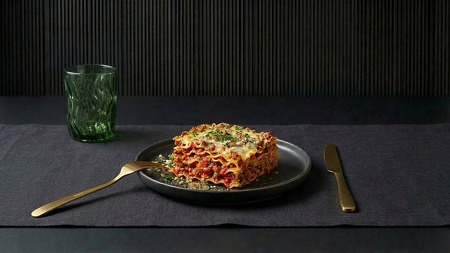 Ein schwarzes Teller mit einer reichhaltigen Lasagne, die Schichten von Nudel, Fleischsoße und geschmolzenem Käse zeigt, steht auf einem dunkelgrauen Tischläufer. Neben dem Teller liegt ein goldener Gabel, und ein grünes Glas steht im Hintergrund.