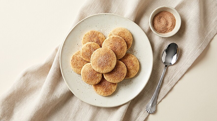 Teller mit mehreren runden Zimtzuckerplätzchen, dicht angeordnet wie eine Blume; daneben kleine Schale mit Zimtzucker und ein Teelöffel auf hellem Leinentuch.