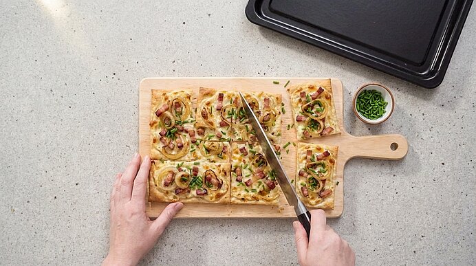 Zwei Hände schneiden einen Flammkuchen mit Speck, Zwiebeln und Schnittlauch auf einem Holzbrett in Rechtecke.
