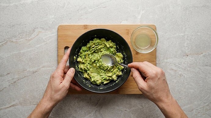 Zwei Hände mischen mit einem Löffel Limettensaft unter die zerdrückte Avocado in einer schwarzen Schüssel.