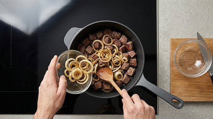 Draufsicht: Hände geben gebratene Zwiebelringe in eine Pfanne mit angebratenen Rindfleischwürfeln; ein Holzlöffel rührt das Gericht auf dem Herd.