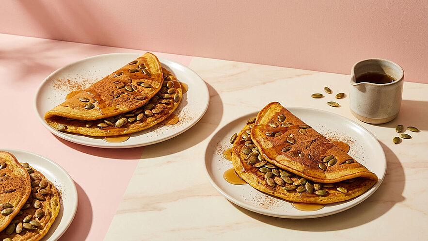Drei Teller mit gefalteten Pfannkuchen, die mit Kürbiskernen und Zimt bestreut sind. Eine Portion hat einen Sirupüberzug. Im Hintergrund steht ein kleiner Behälter mit Sirup und einige Kürbiskerne sind auf der Tischoberfläche verteilt.
