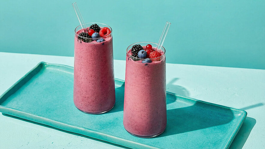 Zwei hohe Gläser mit einem pinken Smoothie stehen auf einem hellblauen Tablett. Die Smoothies sind mit verschiedenen Beeren, darunter Himbeeren, Blaubeeren und Brombeeren, dekoriert und haben jeweils einen Strohhalm. Der Hintergrund ist ein sanftes Blau.