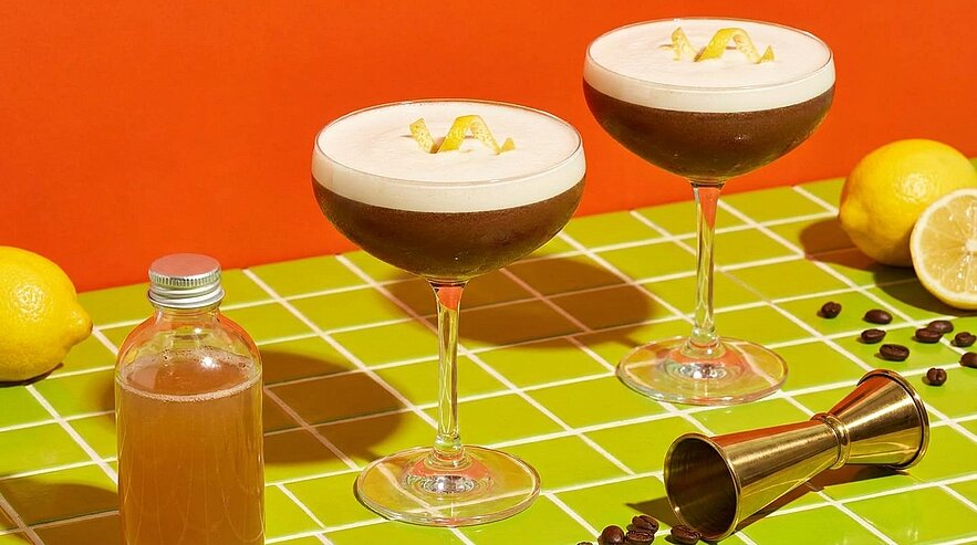 Zwei Espresso‑Martini‑Cocktails in Coupégläsern mit cremiger Schaumkrone, jeweils mit Zitronenzeste garniert, auf grün karierter Oberfläche.