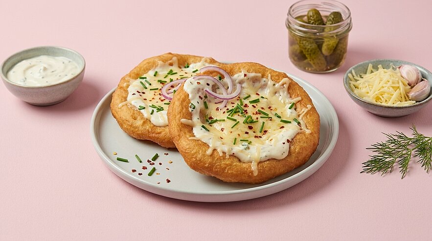 Zwei goldbraun frittierte Langos auf einem Teller, bestrichen mit saurer Sahne und bestreut mit geriebenem Käse, Schnittlauch und roten Zwiebelringen.