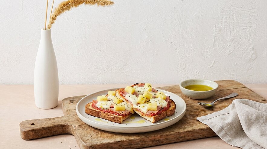 Zwei belegte Toasts auf einem Teller: Schinken, Ananas und geschmolzener Käse, leicht mit schwarzem Pfeffer bestreut, auf einem Holzbrett.