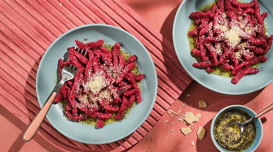 Zwei helle Teller mit purpurroter Rote-Bete-Pasta auf grünem Pesto, mit geriebenem Käse bestreut; in einem Teller liegt eine Gabel. Daneben kleine Schüssel mit Dressing.