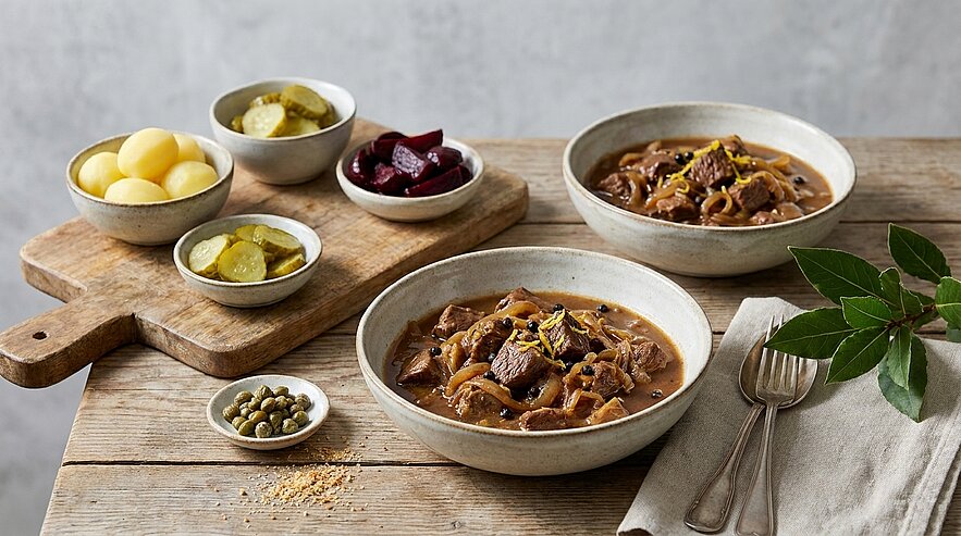 Zwei Schalen Rindergulasch mit Pilzen in dunkler Sauce, dazu kleine Schälchen mit eingelegten Gurken, gekochten Kartoffeln, Roter Bete und Kapern; Besteck und Lorbeerblätter daneben.