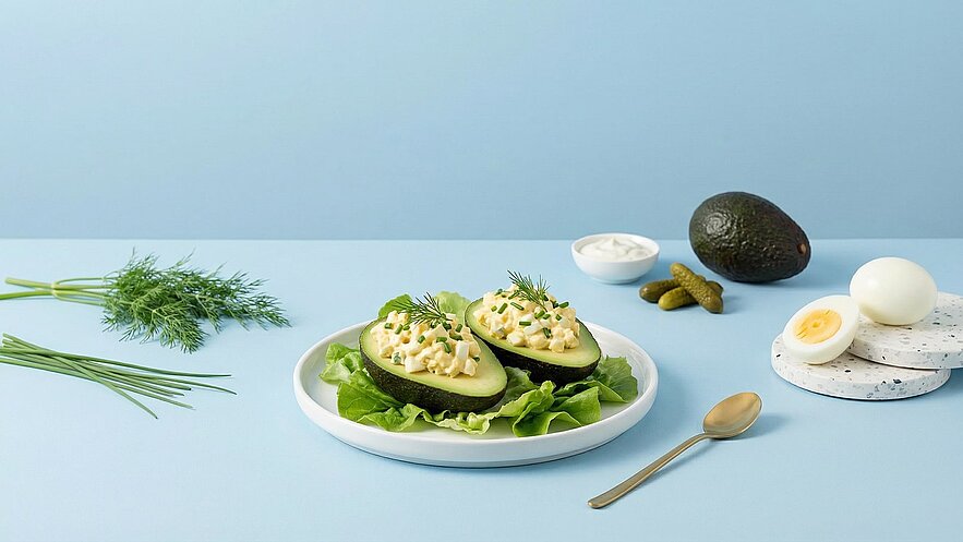 Ein Teller mit zwei halbierten Avocados, gefüllt mit einer Eimischung, steht auf grünem Salat. Daneben liegen frische Kräuter, eingelegte Gurken und ein hartgekochtes Ei auf einem Tisch mit blauer Tischdecke.