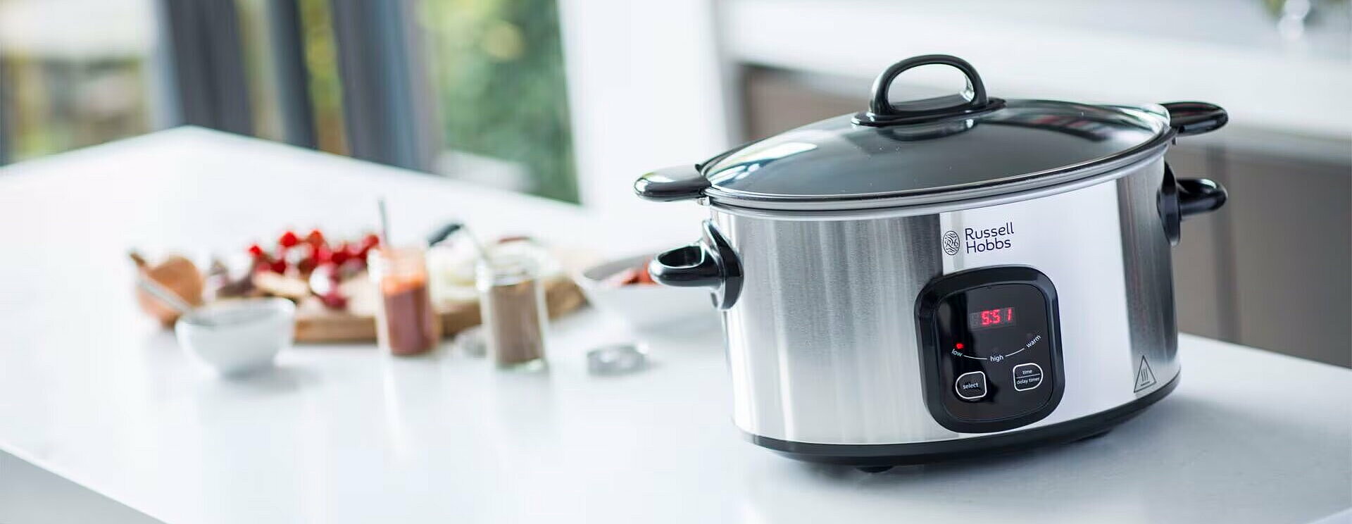 Ein eleganter slow cooker von Russell Hobbs steht auf einer modernen, weißen Küchenarbeitsplatte. Er hat einen schwarzen Deckel und ein Bedienfeld mit digitalen Anzeigen. Im Hintergrund sind verschiedene Lebensmittel und Küchenutensilien zu sehen, die auf das Kochen vorbereiten.
