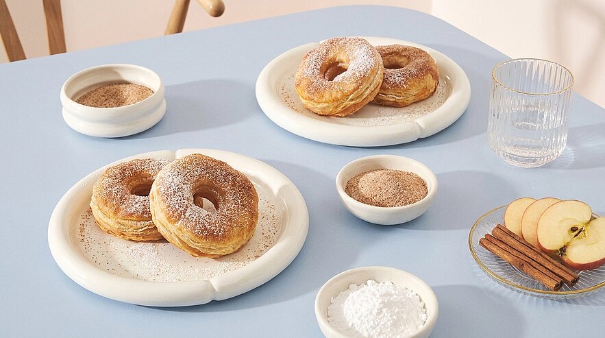 Drei fluffige, mit Puderzucker bestäubte Donuts auf weißen Tellern, begleitet von kleinen Schälchen mit Zucker und Zimt, Apfelscheiben mit Zimtstangen und einem Wasserglas.