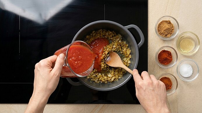 Hände gießen Tomatensauce aus einem Messbecher in einen Topf mit angeschwitzten Zwiebeln und rühren mit einem Holzlöffel auf dem Herd.