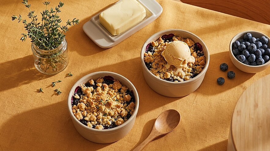Zwei kleine Keramikschalen mit warmem Blaubeer‑Crumble, eine mit einer Kugel Vanilleeis; daneben eine Schale frischer Blaubeeren und zwei Holzlöffel.