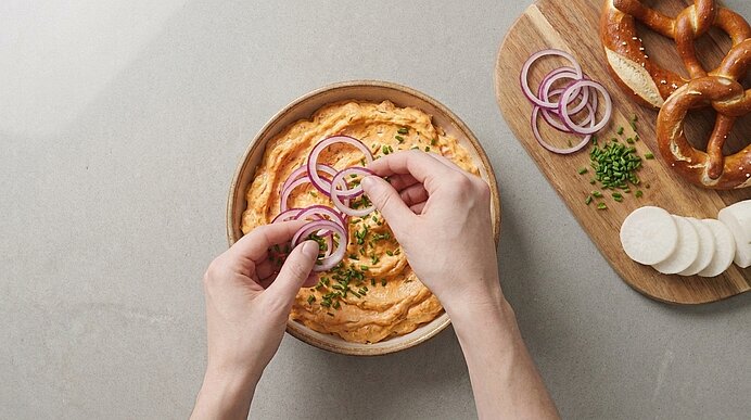 Zwei Hände garnieren eine Schüssel cremigen Käseaufstrich mit roten Zwiebelringen und Schnittlauch; im Hintergrund liegen Brezeln als Beilage.
