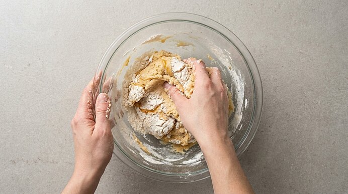 Zwei Hände kneten in einer großen Glasschüssel einen klebrigen Teig aus Mehl und feuchten Zutaten.