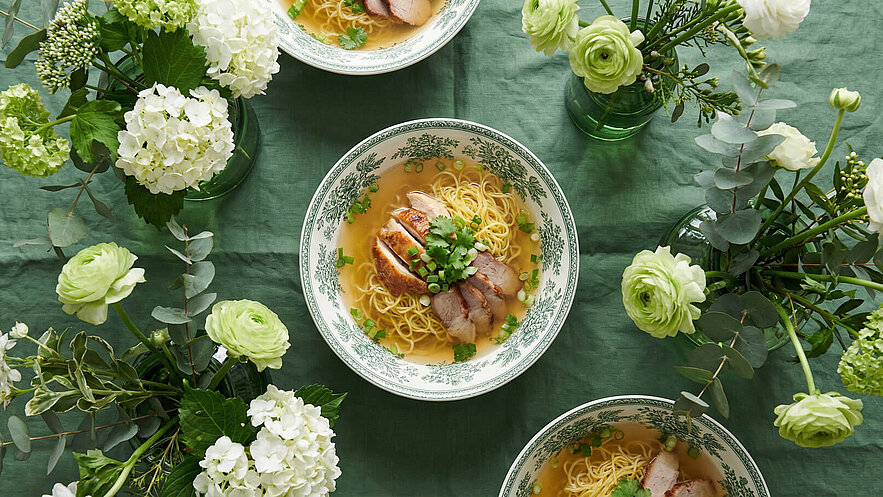Drei Schalen mit Nudelsuppe sind auf einem grünen Tisch gedeckt. Die Suppe enthält dünn geschnittenes Fleisch und ist mit frischem Koriander und Frühlingszwiebeln garniert. Um die Schalen herum stehen Vasen mit weißen und grünen Blumen.