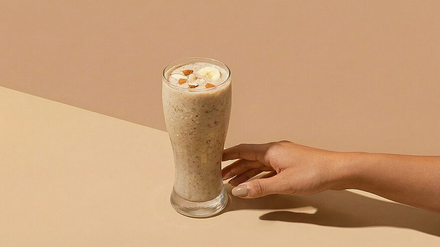 Ein Glas mit cremigem Smoothie steht auf einem Tisch. Der Smoothie ist mit Bananenscheiben und gehackten Mandeln garniert. Eine Hand greift nach dem Glas. Der Hintergrund ist in warmen, neutralen Tönen gehalten.