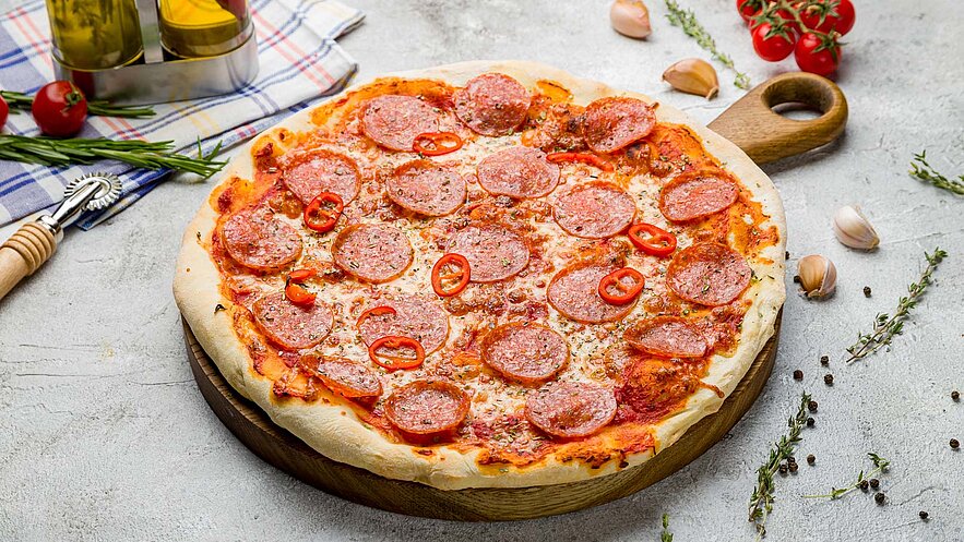 Eine frisch gebackene Pizza liegt auf einem runden Holzbrett. Sie ist mit Tomatensoße, geschmolzenem Käse, zahlreichen Peperoni-Scheiben und roten Chilischoten belegt. Rund um die Pizza sind einige Kräuter, Knoblauchzehen und kleine Tomaten platziert.