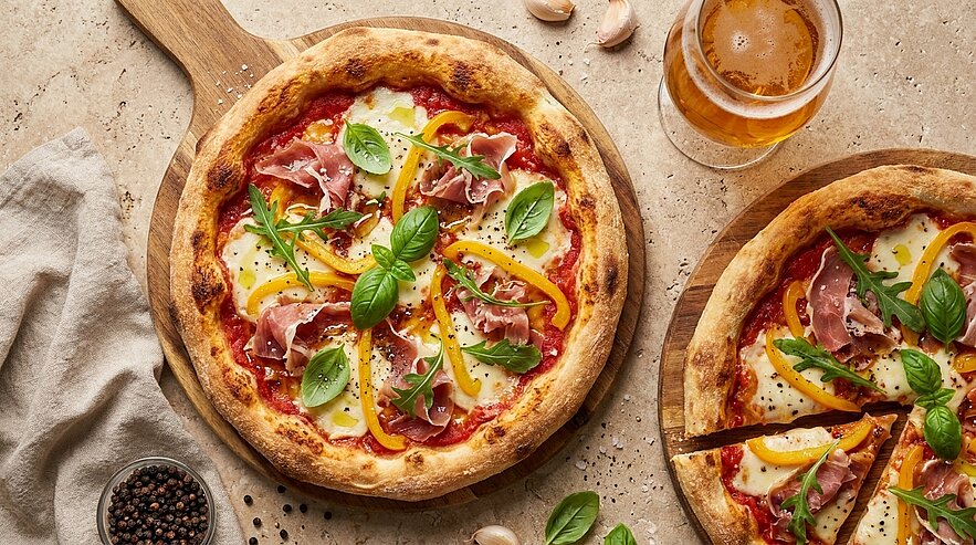 Zwei Pizzen auf Holzbrettchen mit knusprigem Rand, Tomatensauce, geschmolzenem Käse, Parmaschinken, gelber Paprika und frischen Basilikumblättern; eine Pizza ist angeschnitten.