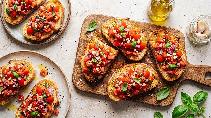 Mehrere geröstete Brotscheiben (Bruschetta) mit gehackten Tomaten, roten Zwiebeln, frischem Basilikum und Olivenöl auf einem Schneidebrett und Tellern angerichtet.