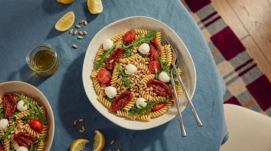 Eine Schüssel Pasta mit Fusilli, frischen Tomaten, Sonnengetrockneten Tomaten, Mozzarella-Kugeln und Rucola. Neben der Schüssel liegen Zitronenscheiben und ein Glas Olivenöl. Der Hintergrund zeigt einen gedeckten Tisch mit einem blauen Tischcloth.