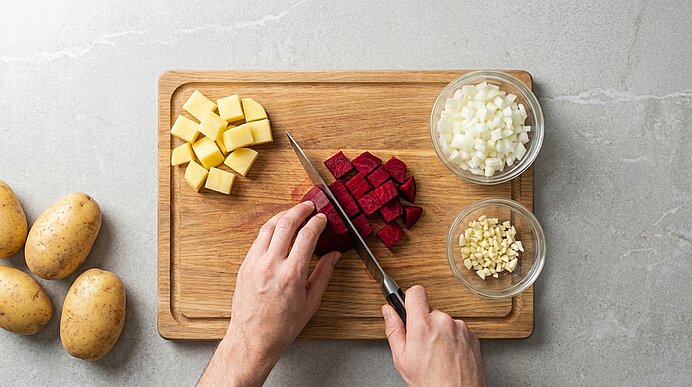Zwei Hände schneiden Rote Bete auf einem Holzbrett, daneben Kartoffelwürfel sowie Schälchen mit Zwiebeln und Knoblauch.