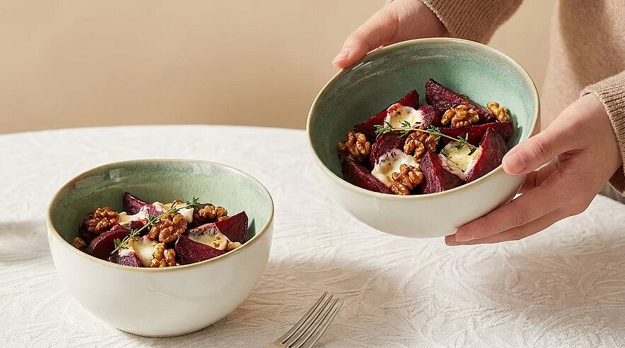 Eine Person hält eine Schüssel mit einer bunten Salatmischung, die Rote Beete, Käse und Walnüsse enthält. Neben ihr steht eine weitere Schüssel mit ähnlichem Salat. Der Tisch ist mit einer weißen Tischdecke gedeckt.