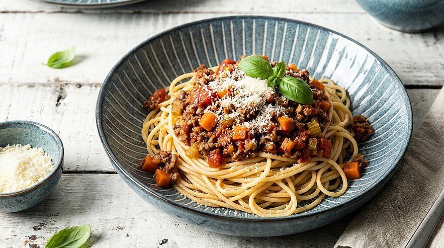 Eine Schüssel mit Spaghetti, die mit einer herzhaften Hackfleischsauce, Karottenwürfeln und frisch geriebenem Käse garniert ist. Dazu liegen einige Basilikumblätter zur Dekoration. Im Hintergrund befindet sich eine kleine Schale mit mehr geriebenem Käse.