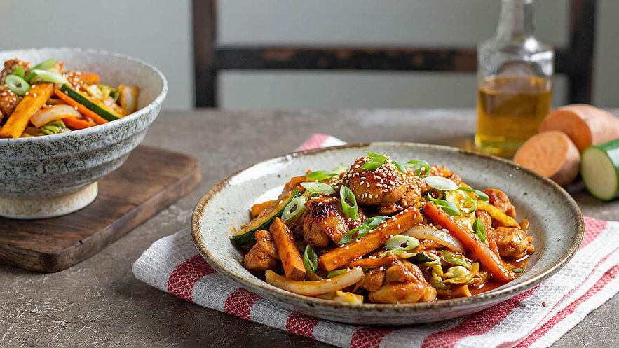 Ein Teller mit gebratenem Hühnchen und Gemüse, darunter Karotten, Zucchini und Frühlingszwiebeln, garniert mit Sesam. Im Hintergrund steht eine Schüssel mit einem ähnlichen Gericht. Das Ganze liegt auf einem gestreiften Tuch.