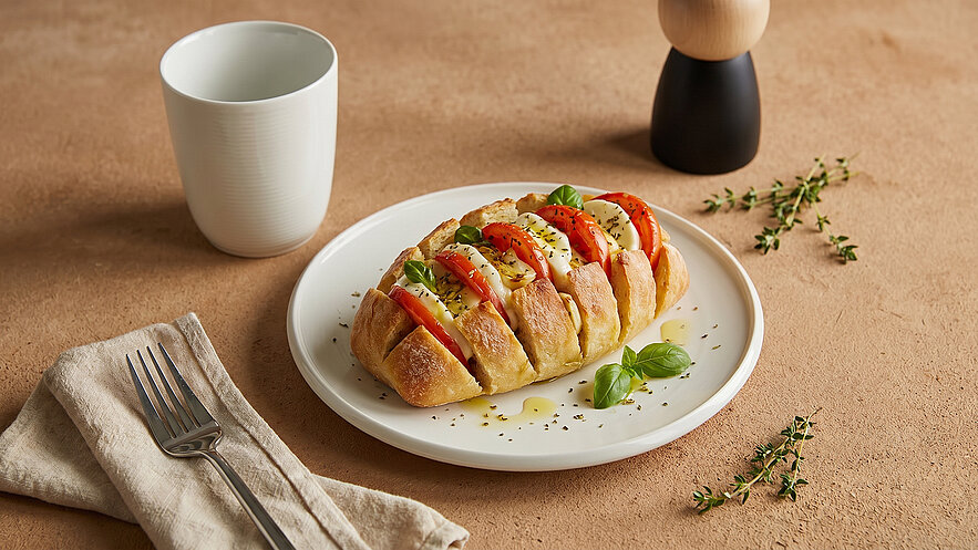 Ein Teller mit einem aufgeschnittenen Brötchen, gefüllt mit Scheiben von Tomaten und Mozzarella. Frische Basilikumblätter und Gewürze sind darauf verteilt. Im Hintergrund steht ein weißer Becher und ein Holzständer. Die Textur des Tellers hebt das Essen hervor.