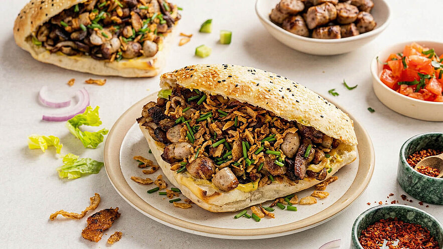 Ein Sandwich aus weichem Brötchen, gefüllt mit gegrilltem Fleisch, Gemüse und frittierten Zwiebeln. Daneben liegen Schalen mit weiteren Fleischstücken sowie eine Schüssel mit gewürfelten Tomaten und Gewürzen. Frische Kräuter verleihen dem Gericht Farbe und Geschmack.