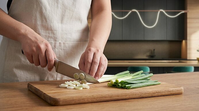 Hände einer Person in Schürze schneiden Frühlingszwiebeln auf einem Holzbrett mit einem Küchenmesser.
