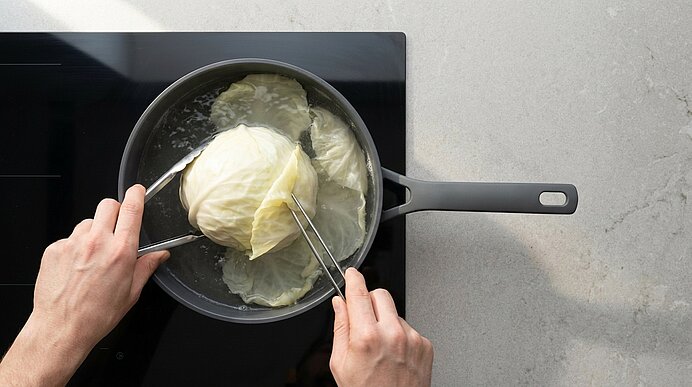 Zwei Hände halten mit Zangen einen ganzen Weißkohl, der in einem Topf mit kochendem Wasser auf einem Herd gegart wird.