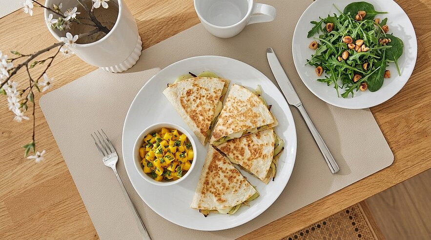 Teller mit goldbraun gebratenen, in Keile geschnittenen Quesadillas, daneben kleine Schale mit Mangosalsa und ein Rucolasalat mit Nüssen; Besteck liegt rechts daneben.
