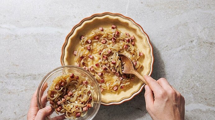 Zwei Hände verteilen mit einem Holzlöffel sautierte Zwiebeln und klein gewürfelten Speck aus einer Glasschüssel in eine Tarteform.